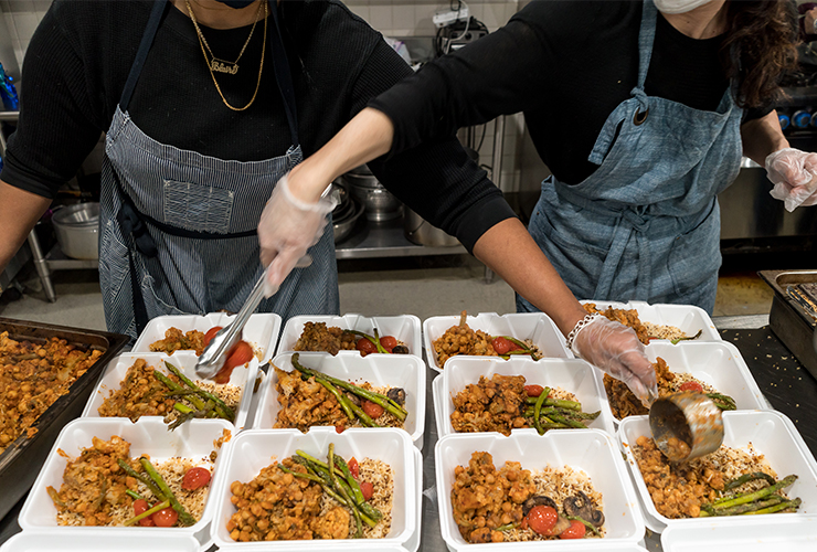 The torsos of two women standing over a table of takeout food containers and scooping food into the containers