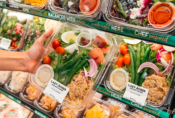A hand holding a packaged salad in front of a shelf of prepared foods